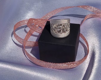 May include: A silver filigree ring with a delicate, intricate design. The ring is displayed on a black square box with a pink ribbon.