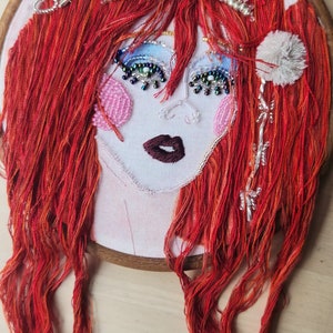 May include: Embroidered portrait of a woman with red hair, wearing a crown of white beads and a white pom-pom on each side of her head. Her eyes are outlined with black thread and decorated with iridescent beads. Her lips are a deep red.