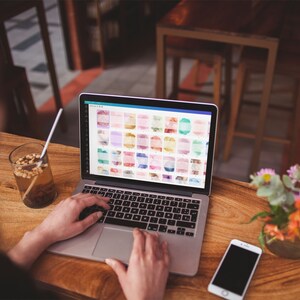 Peut inclure: Un ordinateur portable avec un &eacute;cran affichant une grille de motifs aquarelle color&eacute;s. L'ordinateur portable est ouvert et les mains d'une personne tapent sur le clavier. Un t&eacute;l&eacute;phone portable se trouve sur la table &agrave; c&ocirc;t&eacute; de l'ordinateur portable.