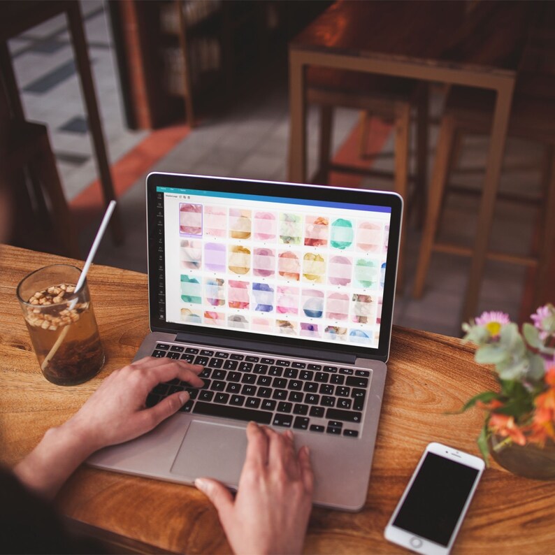 Peut inclure: Un ordinateur portable avec un &eacute;cran affichant une grille de motifs aquarelle color&eacute;s. L'ordinateur portable est ouvert et les mains d'une personne tapent sur le clavier. Un t&eacute;l&eacute;phone portable se trouve sur la table &agrave; c&ocirc;t&eacute; de l'ordinateur portable.