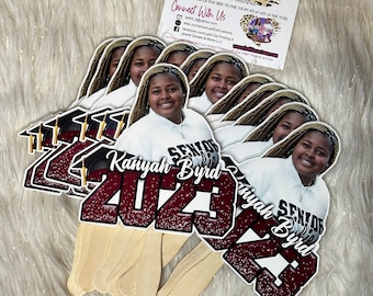 May include: A stack of graduation photo props featuring a young woman in a white shirt. Each prop has the text "Senior 2023" and the name "Kaniyah Byrd" in maroon and white. The props are attached to wooden sticks.
