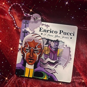 May include: A square keychain featuring a character illustration with the text "Enrico Pucci" and "I Love You Jesus." The design includes a portrait of a person with a star on their head, and a purple and gold color scheme.