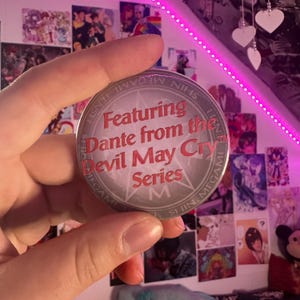 May include: A circular badge with the text "Featuring Dante from the Devil May Cry Series" in red. The badge has a silver border and is held up against a backdrop of posters and a pink light.