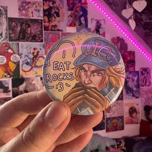 May include: A circular badge with a character illustration and the text "I EAT ROCKS :3". The character is wearing a blue hat with "DIO" on it. The badge has a light-colored background with a cartoon-style design.