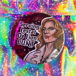 May include: A circular badge featuring a portrait of a person with long blonde hair and blood on their face. The badge has the text "Serving Lestat De Lion Cunt" in white lettering on a maroon background.