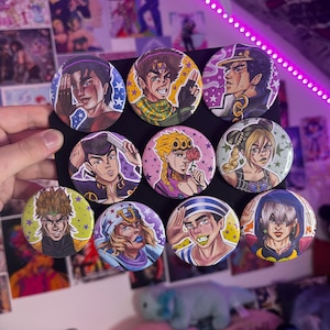 Könnte beinhalten: Eine Sammlung von zehn runden, farbenfrohen Knöpfen mit Illustrationen von Charakteren im Anime-Stil. Jeder Knopf zeigt einen anderen Charakter mit einzigartigen Frisuren und Outfits. Die Knöpfe werden vor einem schwarzen Hintergrund gehalten, mit einer Hand im Bild.