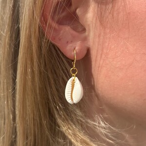 May include: A gold hoop earring featuring a small, white seashell pendant. The seashell is oval and has a ridged texture. The earring is attached to an ear, with blonde hair in the background. The earring is a simple, elegant design.