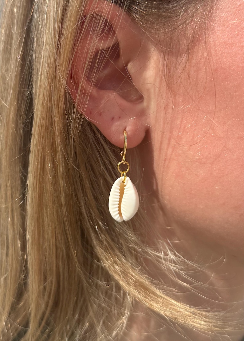 May include: A gold hoop earring featuring a small, white seashell pendant. The seashell is oval and has a ridged texture. The earring is attached to an ear, with blonde hair in the background. The earring is a simple, elegant design.