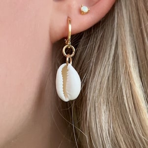 May include: A gold hoop earring with a small, white seashell pendant. The earring is attached to a pierced ear with other gold and gemstone earrings. The background shows blonde hair.