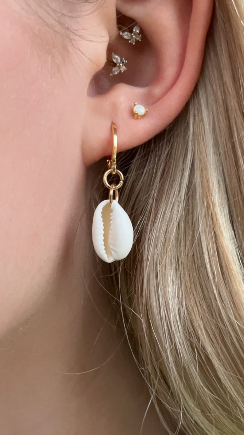 May include: A gold hoop earring with a small, white seashell pendant. The earring is attached to a pierced ear with other gold and gemstone earrings. The background shows blonde hair.