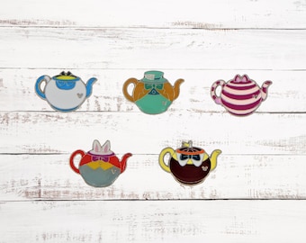 May include: Five enamel pins shaped like teapots, each featuring a different character from Alice in Wonderland. The characters are the Mad Hatter, the March Hare, the Queen of Hearts, the Cheshire Cat, and the White Rabbit.