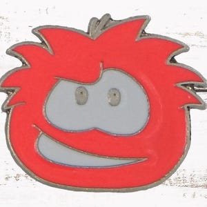 May include: A red, cartoon-style pin featuring a smiling character with a red body, gray eyes, and a white mouth. The pin has a silver-colored outline and is set against a white wooden background.