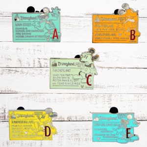 May include: Five Disneyland ticket pins featuring Mickey Mouse. Each pin is a different color and features a different attraction. The pins are labeled A through E.
