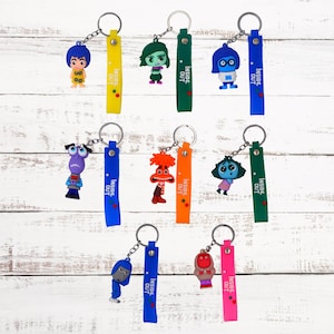 May include: Nine colorful keychains featuring characters from the movie Inside Out. Each keychain has a different character, including Joy, Sadness, Anger, Fear, Disgust, Bing Bong, and a blue character with a crown. The keychains are all attached to a silver key ring and have a blue, green, yellow, orange, or red strap with the words "Inside Out" printed on them.