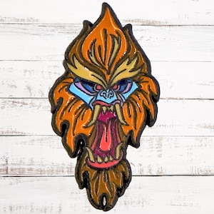 May include: A colourful enamel pin depicting a cartoon monkey with orange fur, blue eyes, and a large, open mouth with sharp teeth. The monkey's face is detailed with a yellow brow and a brown beard.