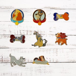 May include: A collection of seven enamel pins featuring characters from the Disney movie Lady and the Tramp. The pins are in various shapes and sizes, including a bone, a dog bed, and a dog collar. The pins are all colorful and feature detailed artwork.