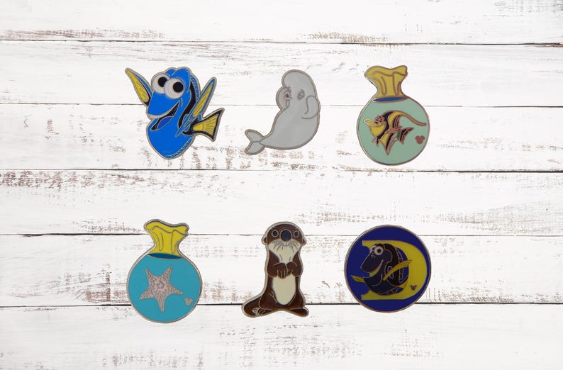 Puede incluir: Seis pines de esmalte con personajes de la pel&iacute;cula animada Buscando a Nemo. Los pines est&aacute;n sobre un fondo de madera blanca. Los pines incluyen a Dory, un pez cirujano azul, una ballena beluga blanca, un pez &aacute;ngel amarillo y naranja en una bolsa azul, una nutria marina marr&oacute;n y un pez payaso azul y amarillo en una bolsa azul.