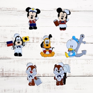 May include: A collection of cartoon character figurines, including Mickey Mouse, Minnie Mouse, Goofy, Pluto, Donald Duck, and Chip and Dale, all dressed in nautical-themed outfits. The figurines are set against a white wooden background.