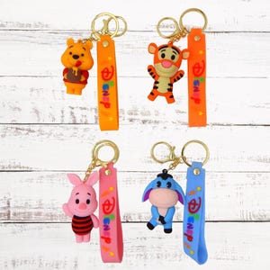 Könnte beinhalten: Vier bunte Schlüsselanhänger mit Disney-Charakteren. Die Schlüsselanhänger sind an orange, rosa, blauen und gelben Bändern befestigt, auf denen das Wort "Disney" steht. Die Figuren sind Winnie Puuh, Tigger, Ferkel und Esel.