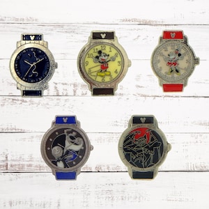 May include: Six decorative watch pins with various designs. The watches have silver-colored cases and straps in blue, red, and black. The watch faces feature cartoon characters and abstract designs. The pins are displayed on a white wooden surface.