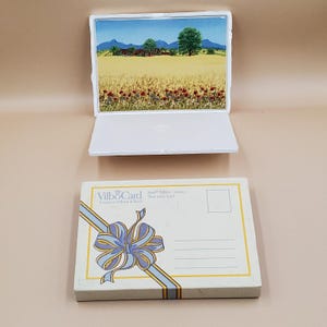 Puede incluir: Un marco de fotos blanco con una pintura de paisaje de un campo de trigo, amapolas rojas y un cielo azul. Una postal con un diseño de cinta azul y amarilla y el texto "Vilbo Card" debajo.