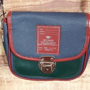 May include: A navy blue and green crossbody bag with red trim. The bag features a rectangular red patch with the text "SPLENDID CREATOR LEADER OF YEAR 2000 SINCE 1995". It has a silver clasp and a silver chain strap.