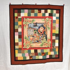 May include: A patchwork quilt with a central image of a scarecrow standing in front of a sign that reads "Harvest Hill Antiques and More", surrounded by sunflowers and apples. The quilt is made of various colors and patterns, including red, green, yellow, and brown.