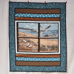 May include: A quilted wall hanging featuring a coastal scene with seagulls. The central image depicts a beach with seagulls, framed by a window design. The quilt has a blue and brown border with a patterned design.