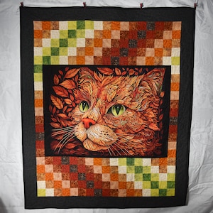 May include: A handmade quilt featuring a detailed cat portrait. The cat's face is rendered in shades of orange and red, surrounded by leaf-like patterns. The quilt has a patchwork border in autumnal colors and a dark gray frame.