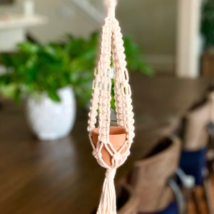 Peut inclure: Un porte-plante en macramé beige avec un pot en terre cuite suspendu. Le porte-plante a un pompon en bas.
