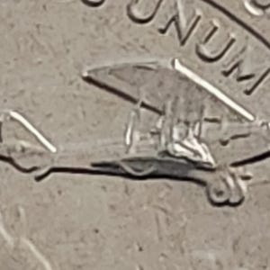 Może przedstawiać: Zbliżenie srebrnej monety z małym wzorem samolotu i napisem "E PLURIBUS UNUM".