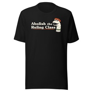 May include: A black short-sleeved t-shirt featuring a graphic of a white sock puppet wearing a red beret. The text "Abolish the Ruling Class" is printed in white and orange lettering next to the puppet, with a horizontal orange line underneath.