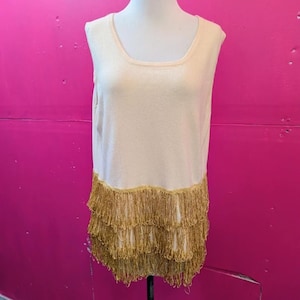 May include: Cream-colored sleeveless top with a scoop neckline and a gold fringe hem. The fringe is made of multiple layers of thin, gold strands. The top is displayed on a white mannequin against a bright pink background.