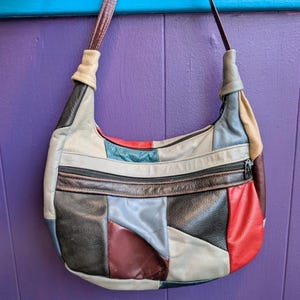 May include: A patchwork leather shoulder bag with a curved shape. The bag features a variety of colors, including red, black, gray, and blue. It has a zipper closure and a brown strap.