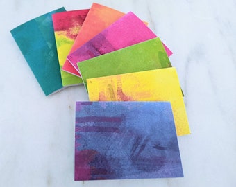 Op de afbeelding: Een set van zes kleine notitieboekjes met kleurrijke aquarelhoezen. De hoezen hebben abstracte penseelstreken in tinten blauw, groen, geel, roze, oranje en turquoise.