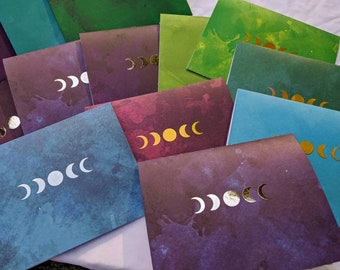 May include: A stack of cards with watercolor backgrounds and gold and silver moon phases printed on them.
