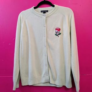 Könnte beinhalten: Hellgrüne Strickjacke mit Rundhalsausschnitt und perlenartigen Knöpfen. Der Pullover hat ein gesticktes Design eines Schädels mit einer roten Rose auf der linken Brust. Der Markenname "Design Original" ist sichtbar.