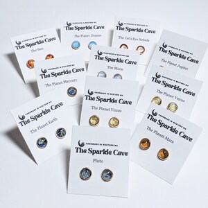 May include: A set of 10 pairs of handmade earrings featuring different planets and celestial bodies. Each pair is displayed on a white card with the name of the celestial body and the words "The Sparkle Cave".