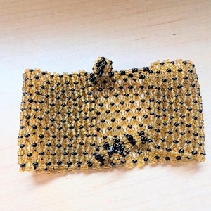 Könnte beinhalten: Ein rechteckiges Manschettenarmband aus einem Gitter kleiner, durchscheinender bernsteinfarbener Perlen und blickdichter schwarzer Perlen. Die gewebte Perlenarbeit zeigt ein sich wiederholendes Rautenmuster mit einem kleinen Perlenverschluss.