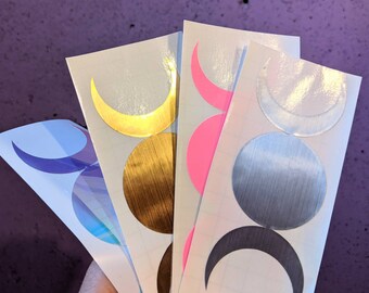 Puede incluir: Cuatro hojas de papel adhesivo blanco con un diseño de triple luna. Cada hoja presenta un color y acabado diferente: dorado, rosa, plateado y azul iridiscente.