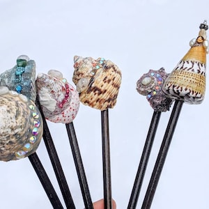 May include: Five decorative sticks with seashells attached to the top. The shells are decorated with iridescent rhinestones and pearls. The sticks are black and the shells are various colors, including brown, white, blue, and pink.