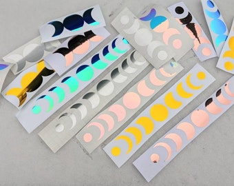 May include: Assortment of moon phase stickers in various colors, including silver, gold, pink, blue, and iridescent. The stickers are arranged on white sheets of paper.