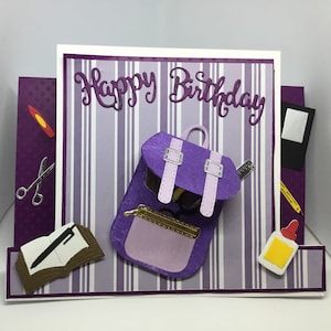 May include: Purple birthday card with a purple backpack, a book, a pencil, scissors, a ruler, glue, and a crayon. The card says "Happy Birthday".