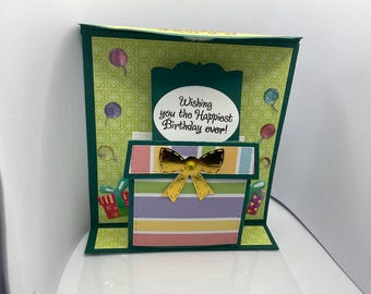 May include: A green and white card with a pop-up box that says "Wishing you the Happiest Birthday ever!" The box is decorated with a gold bow and colorful stripes.