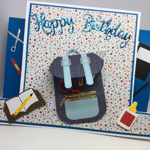 May include: A blue and white backpack with a zippered pocket and a yellow zipper pull. The backpack is filled with school supplies, including a pencil, scissors, and a ruler. The background is a white card with colorful polka dots and the words "Happy Birthday" in blue glitter.