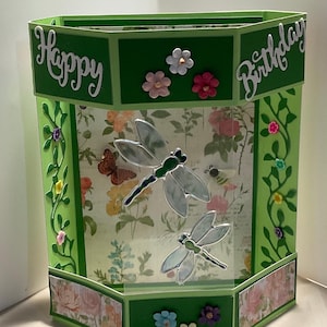 May include: A hexagonal green paper card featuring the text Happy Birthday in white script. The center panel displays a floral background with two translucent dragonfly cutouts, while the side panels are decorated with green vine die-cuts and small colorful flower embellishments.