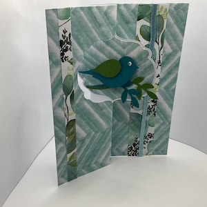 May include: A teal blue bird with green wings perched on a branch, cut out of paper and layered on a green and white patterned card.