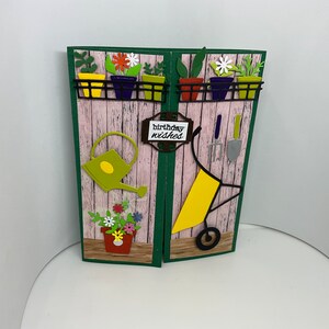 May include: A handmade greeting card with a gardening theme. The card features a pink wooden background with green trim. There are cut-out images of flowers, plants, a watering can, a wheelbarrow, and gardening tools. The card says "birthday wishes" on a brown sign.