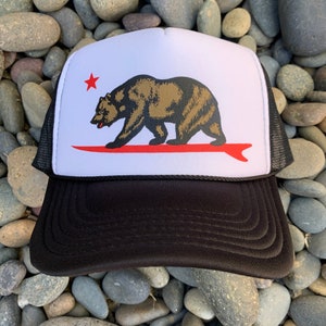 Puede incluir: Gorra de camionero en blanco y negro con la bandera del estado de California. El diseño de la bandera incluye un oso pardo sobre una tabla de surf roja, una estrella roja y un fondo blanco. La gorra tiene una visera negra y una parte trasera de malla.