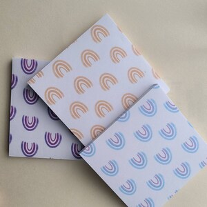 May include: Three square sticky note pads with different rainbow patterns. The top pad has a light orange rainbow pattern, the middle pad has a purple rainbow pattern, and the bottom pad has a light blue rainbow pattern.